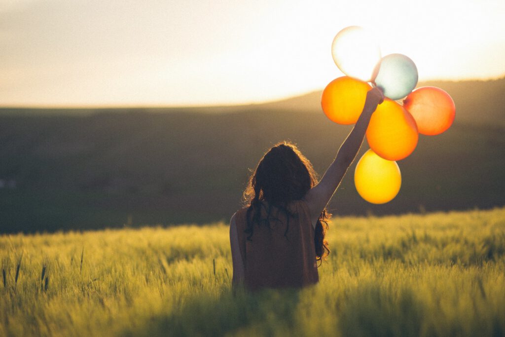 how to find happiness again: woman holding up a bouquet of baloons in a field at sunset