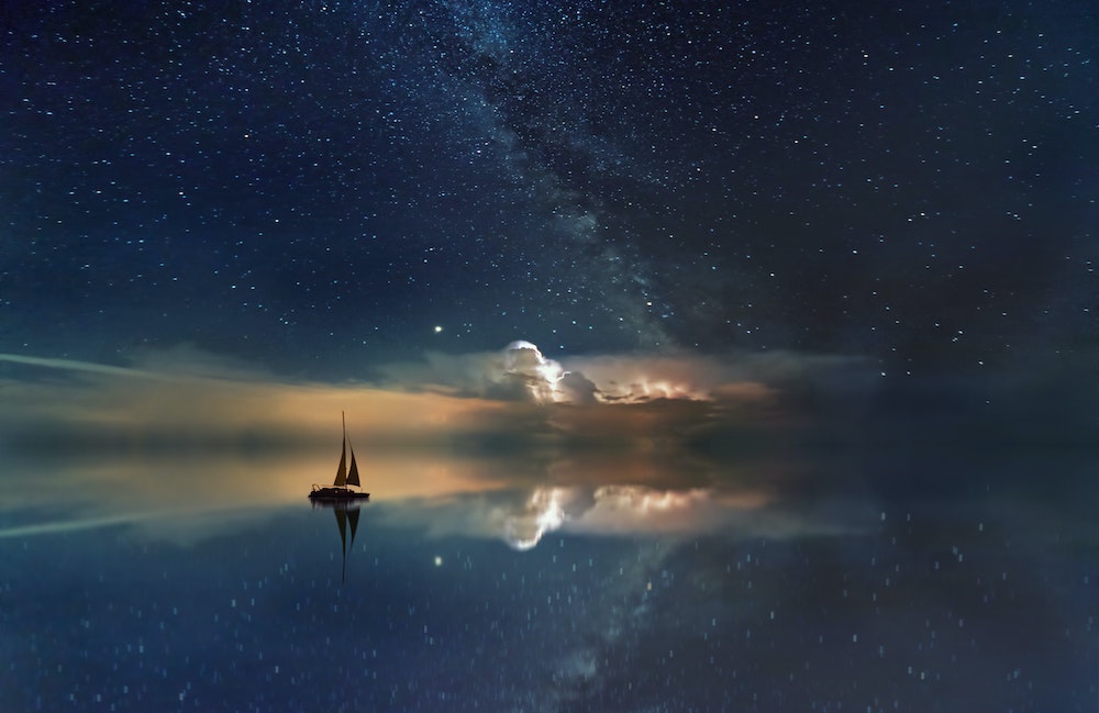 dream-like looking ocean with sailboat and the moon under a starry sky